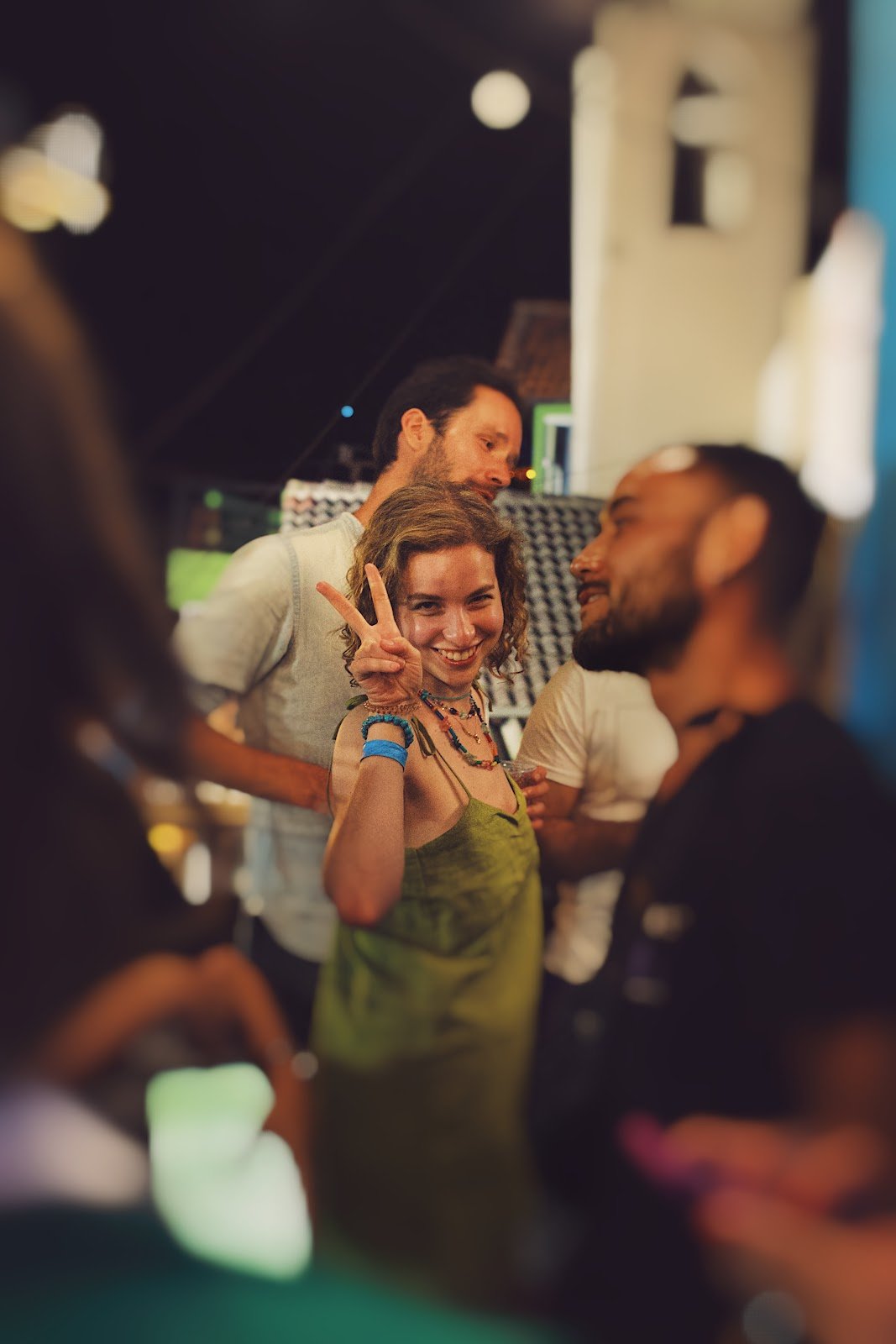 Pessoas aproveitando uma noite de festas no hostel, foco em uma mulher de cabelos curtos, cacheados e loiros, com um vestido verde fazendo sinal de "amor e paz" com os dedos. 